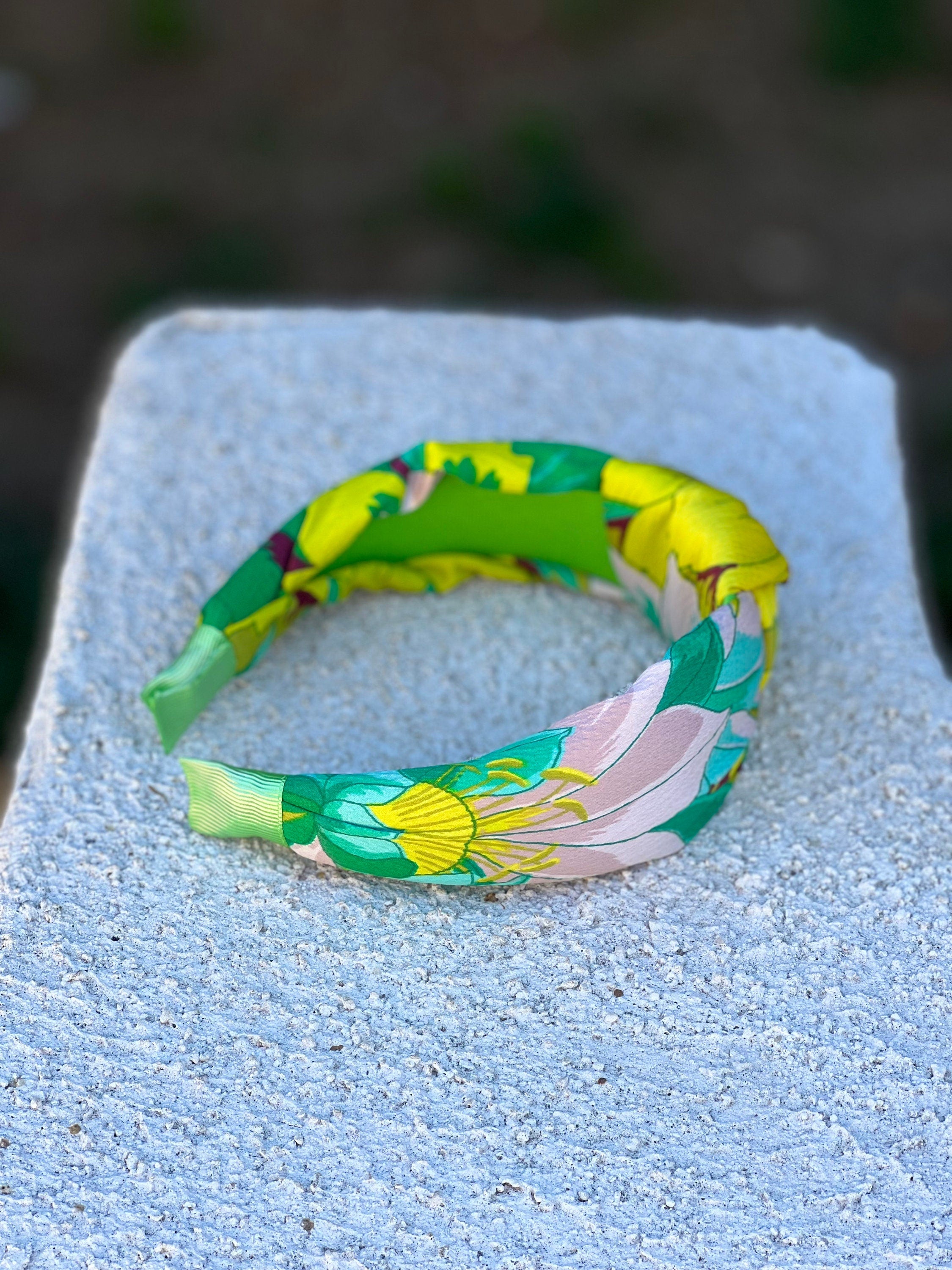 The green and yellow flower pattern knotted headband rests on a wooden table, next to a vase of fresh flowers.